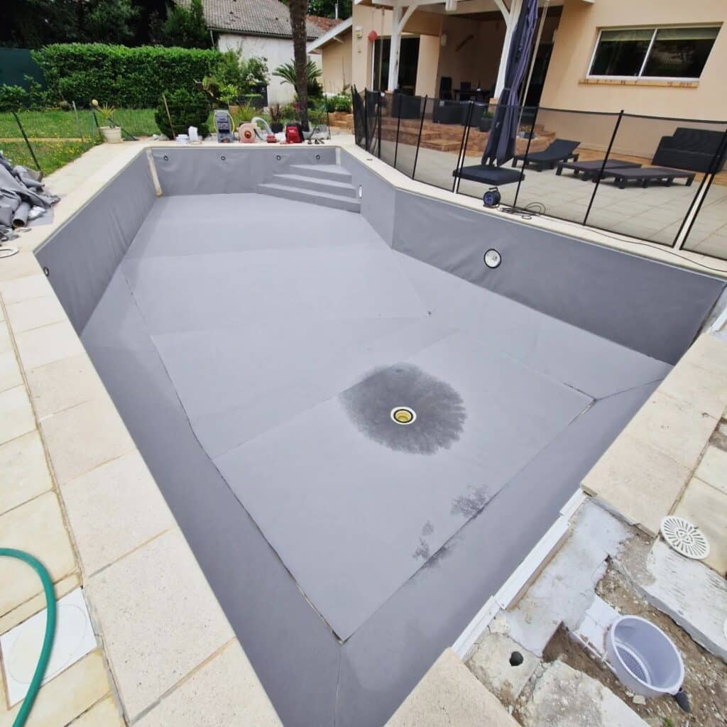 Piscine vide avec revêtement de protection gris et escalier intégré, prête pour l'installation du liner. Entourage pavé, maison et terrasse sécurisée.