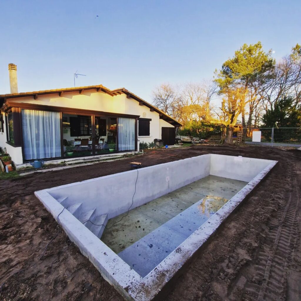 Coque de piscine rectangulaire en béton brute, en construction, devant une maison moderne au toit de tuiles. Le terrain est en terre battue.