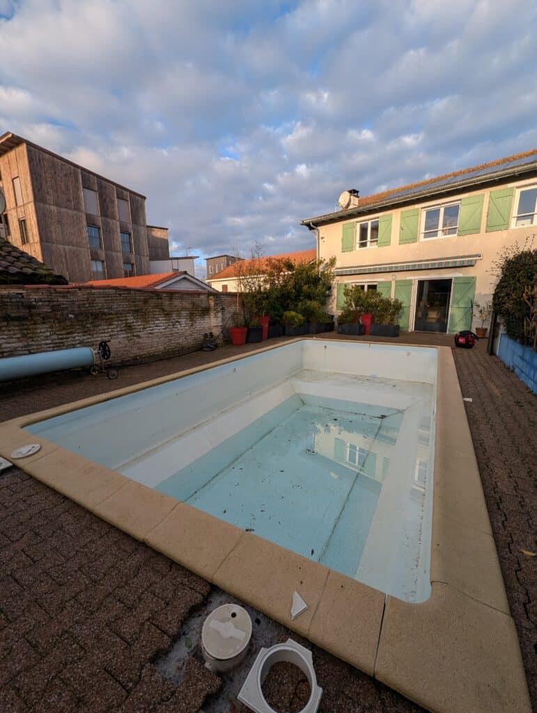 Piscine de jardin partiellement vidée et sale avec débris, entourée de dalles abîmées. Maison aux volets verts et bâtiment moderne sous ciel nuageux.