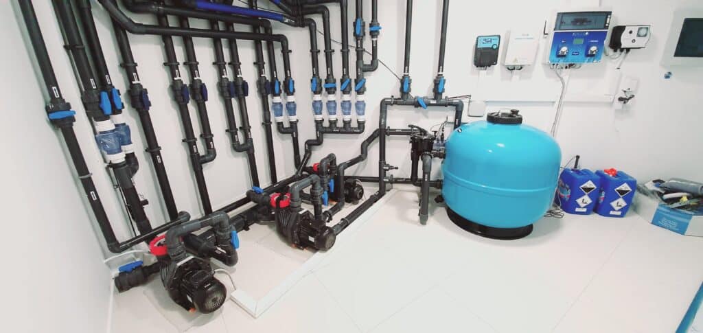 Salle technique piscine : filtration et traitement de l'eau Salle technique de piscine avec réseau de tuyaux noirs, vannes bleues, trois pompes, un filtre à sable bleu et deux bidons UN 1791.