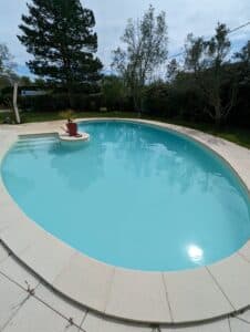 Piscine ovale aux eaux bleu turquoise, bordée de margelles claires, avec une plante en pot rouge près des marches d'accès, entourée de verdure.