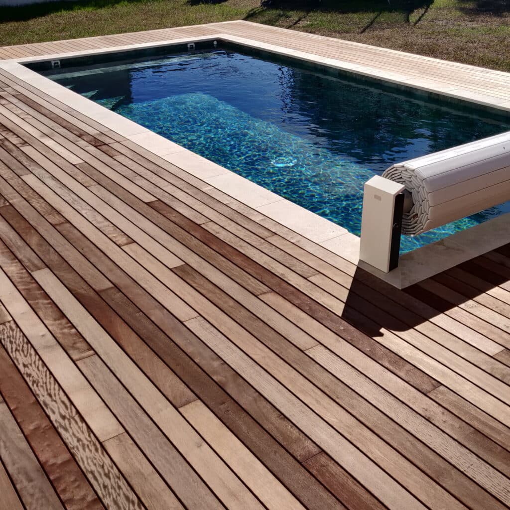 Terrasse en bois moderne bordant une piscine rectangulaire aux eaux bleues, avec volet roulant partiellement enroulé.